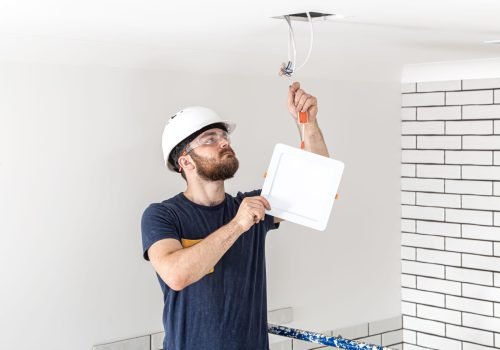 electrician-builder-with-beard-worker-white-helmet-work-installation-lamps-height-professional-overalls-with-drill-repair-site (1)